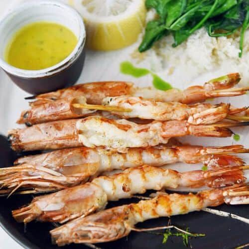 Fresh cooked shrimp with lemon, greens, and dipping sauce on a black plate.